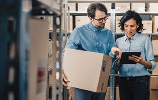 Female Inventory Manager Shows Digital Tablet Information to a Worker Holding Cardboard Box, They Talk and Do Work. In the Background Stock of Parcels with Products Ready for Shipment.