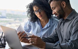 Multiethnic male indian mentor and female African American intern sitting at desk with laptop doing paperwork together discussing project financial report. Corporate business collaboration concept.